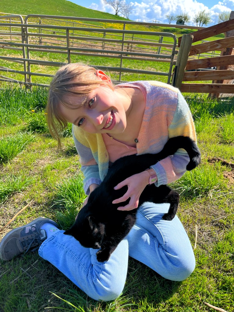 Emily is smiling with blonde bangs with her hair pulled back. She has a colorful cardigan and is holding a black cat in her lap. Behind her is a farm-like horizon.