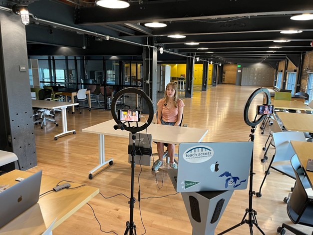 Emily sits at a table in an office room while being filmed on a phone camera.